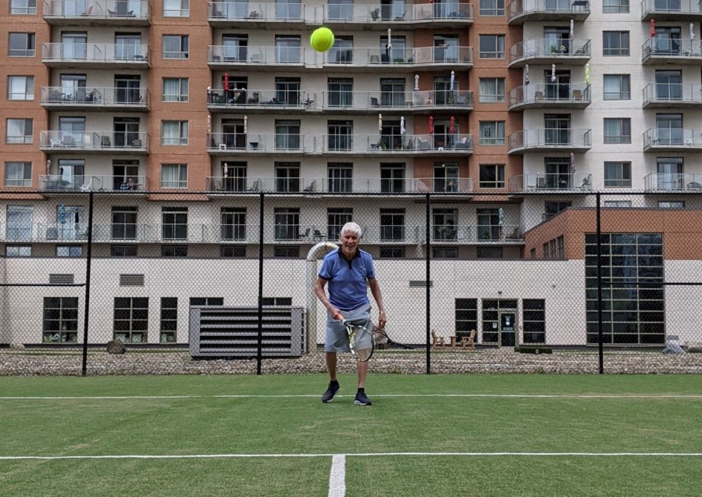 A senior man plays tennis and smiles to the camera 
