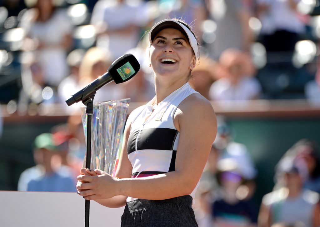 Bianca Andreescu smiles in front of a microphone under the sun, with Indian Wells trophy in the back