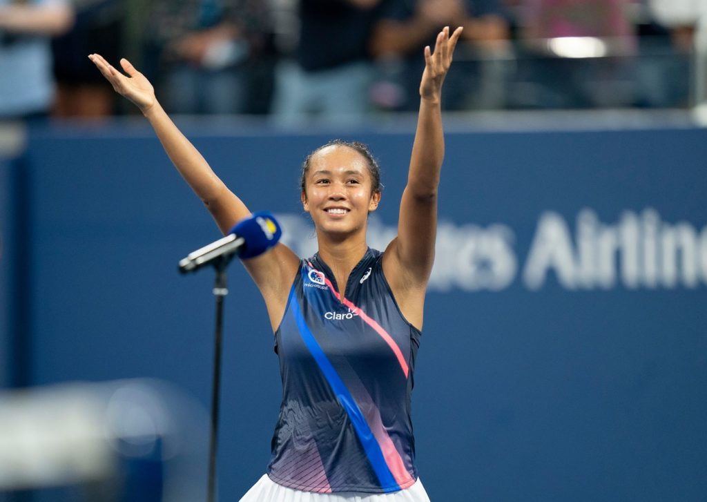 Leylah Fernandez standing in front of a mic on court and lifting her arms and smiling