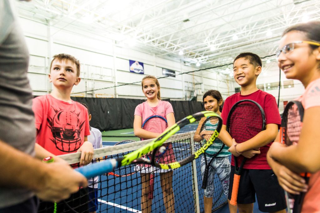 Group of kids holding tennis rackets looking at a coach and smiling
