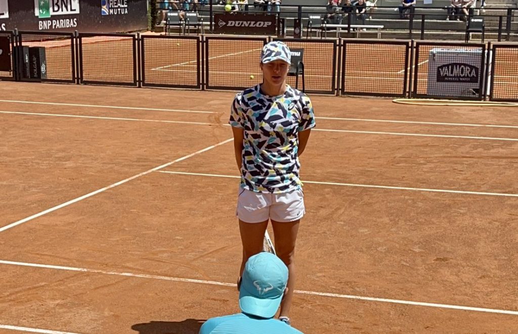 Iga Swiatek stands in front of Rafael Nadal (seated, back to camera)