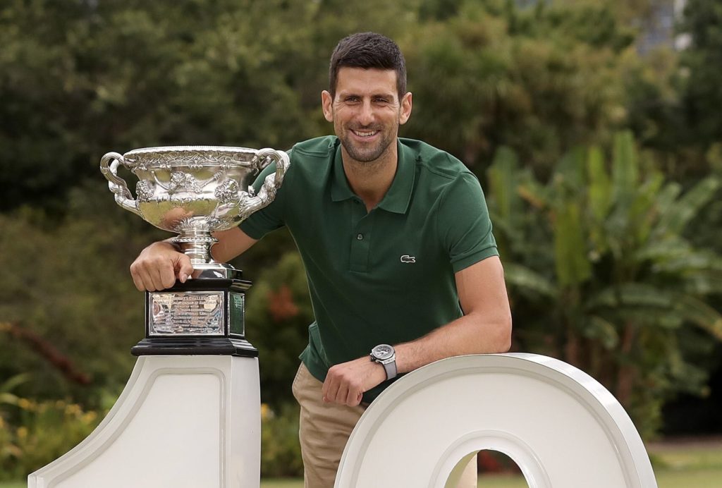Novak Djokovic poses with the Australian Open trophy while leaning on a giant "10" sign