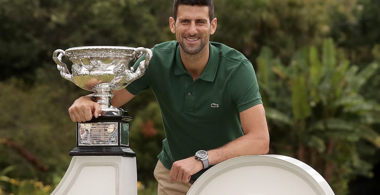 Novak Djokovic poses with the Australian Open trophy while leaning on a giant "10" sign