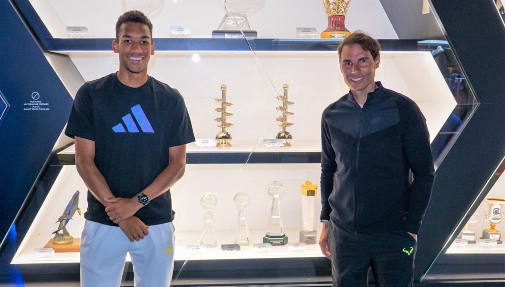 Felix Auger-Aliassime stands next to Rafael Nadal in front of a trophy case.