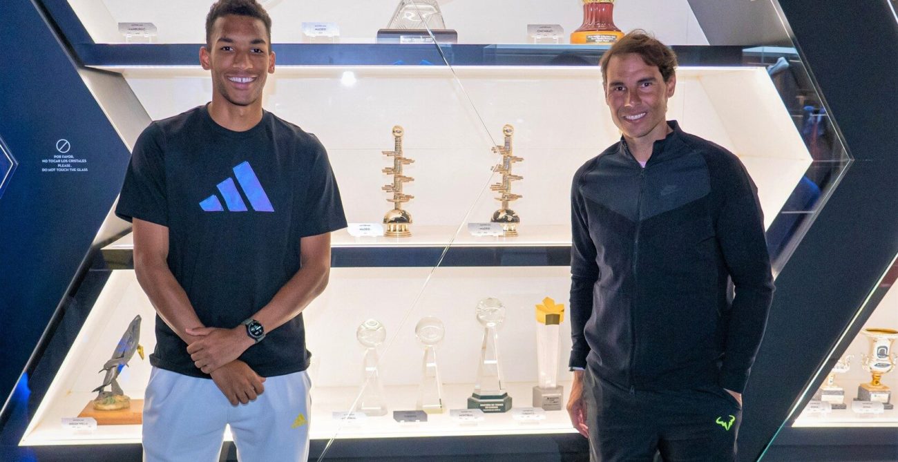 Felix Auger-Aliassime stands next to Rafael Nadal in front of a trophy case.