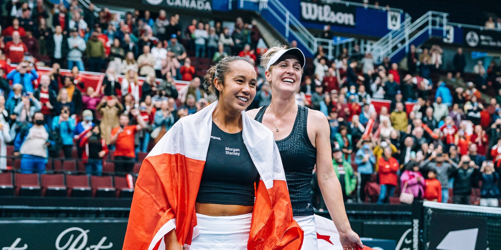 Leylah Fernandez (left) and Gabriela Dabrowski smile on court holding Canadian flags.