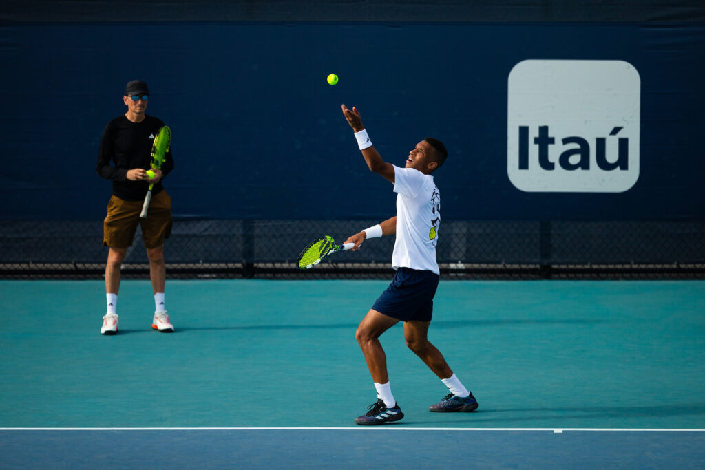 Felix Auger-Aliassime tosses a ball up to serve as Fred Fontang stands by the back wall and observes.