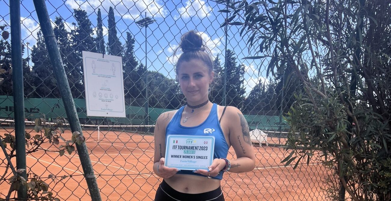 Andreea Prisacariu holds up a winners plate in front of a clay court.
