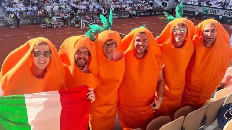A bunch of men in carrot suits holding an Italian flag stand above a tennis court.
