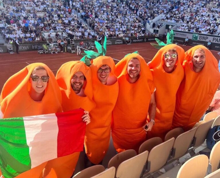 A bunch of men in carrot suits holding an Italian flag stand above a tennis court.